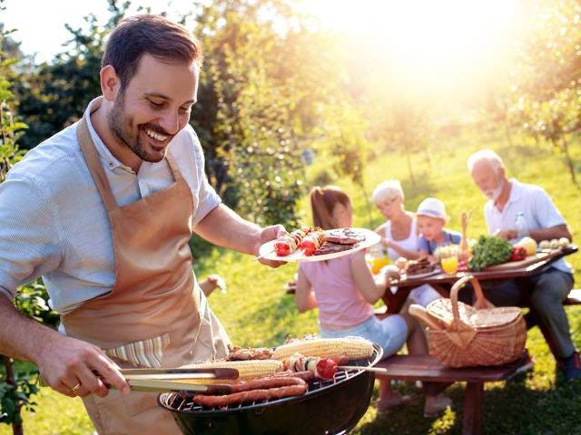 Man staat achter barbecue worstjes te grillen terwijl het gezin buiten aan tafel geniet van het eten